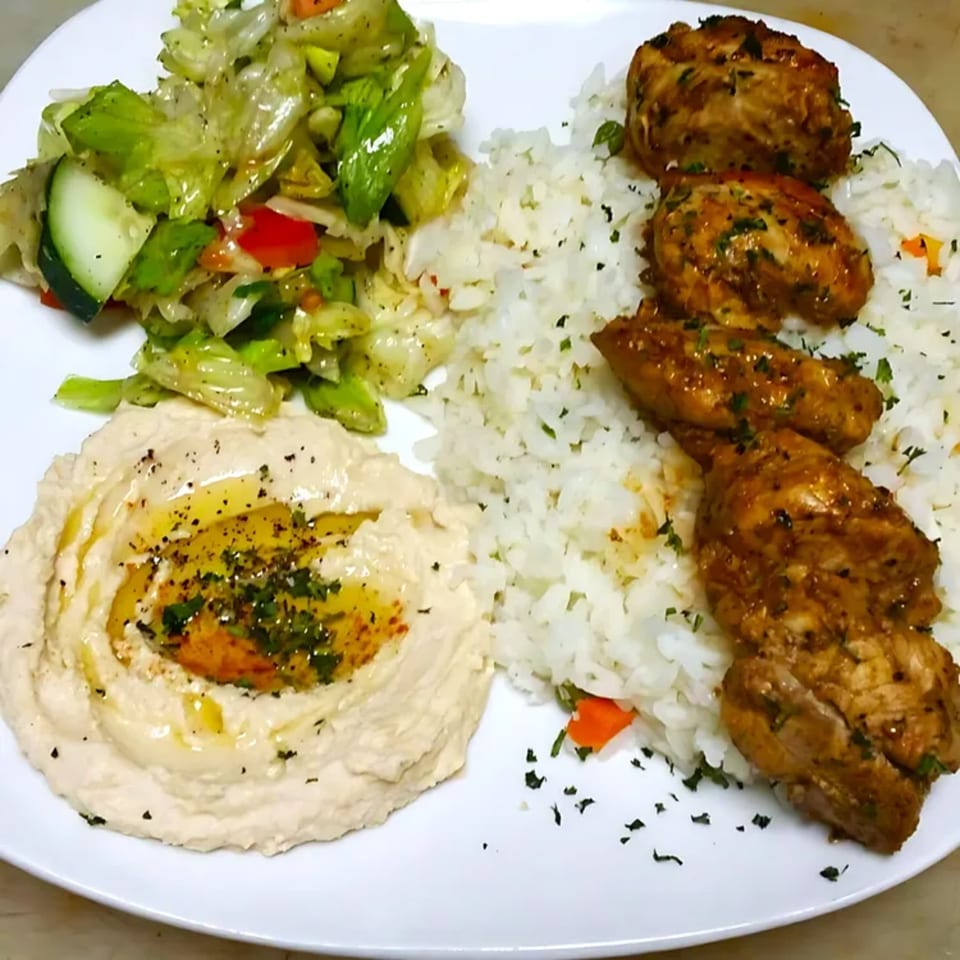 Mediterranean plate with chicken, rice, hummus drizzled with olive oil, and a side salad.