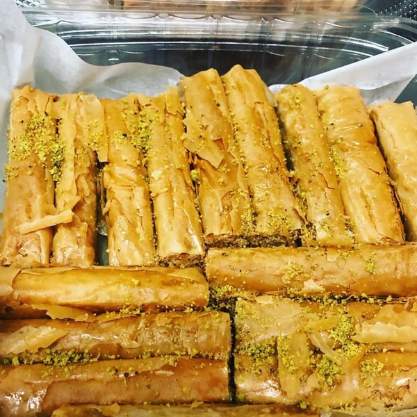 Close-up of golden baklava rolls, sprinkled with green pistachios, in a clear plastic container.