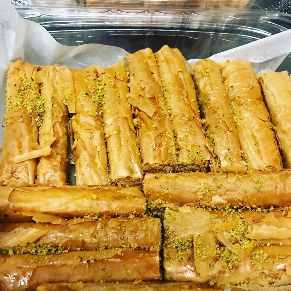 Close-up of golden baklava rolls, sprinkled with green pistachios, in a clear plastic container.