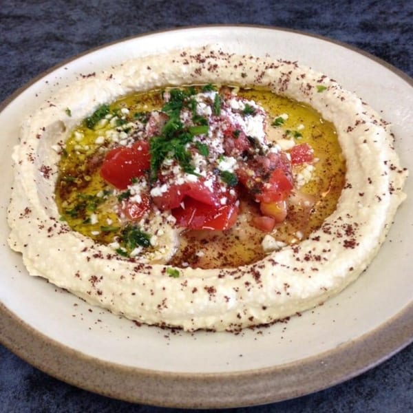 Creamy hummus topped with olive oil, diced tomatoes, feta cheese, and parsley on a plate.